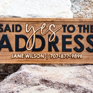 May include: A rectangular wooden sign with the phrase "SAID YES TO THE ADDRESS" in a mix of fonts and colors. The sign includes a name and phone number. The sign is brown with black and light brown lettering.