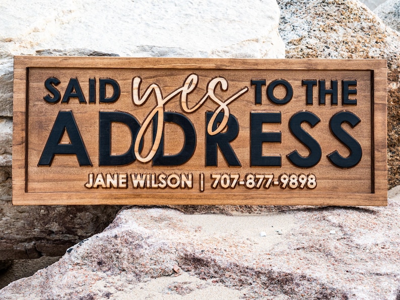 May include: A rectangular wooden sign with the phrase "SAID YES TO THE ADDRESS" in a mix of fonts and colors. The sign includes a name and phone number. The sign is brown with black and light brown lettering.