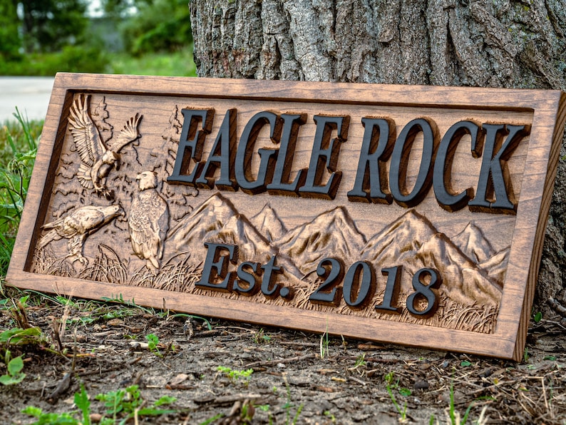 May include: A wooden sign with the text "Eagle Rock Est. 2018" carved into it. The sign features a carved image of two eagles and a mountain range.