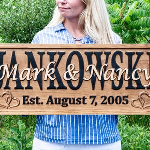 May include: A wooden sign featuring the names "Mark & Nancy" in a decorative font, with "Jankowski" above. The sign includes the date "Est. August 7, 2005" and heart-shaped designs. The sign is brown with black and light brown lettering.