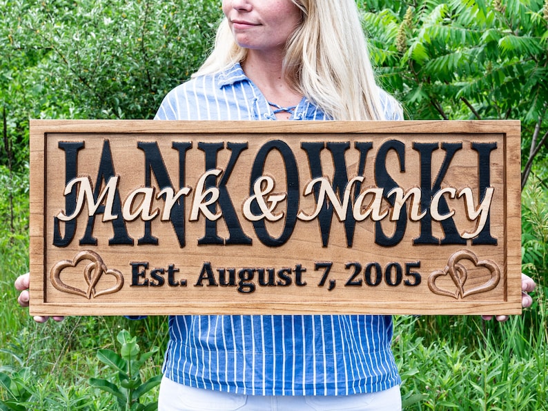 May include: A wooden sign featuring the names "Mark & Nancy" in a decorative font, with "Jankowski" above. The sign includes the date "Est. August 7, 2005" and heart-shaped designs. The sign is brown with black and light brown lettering.