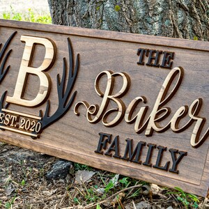 May include: A wooden sign with the text "The Baker Family" in a decorative script font. The sign also features a large "B" initial, a pair of antlers, and the text "EST. 2020".