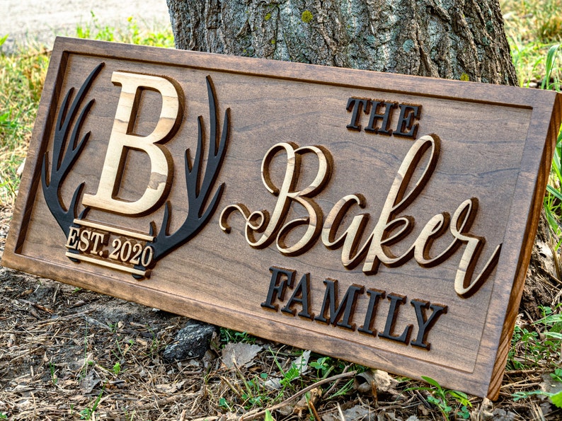 May include: A wooden sign with the text "The Baker Family" in a decorative script font. The sign also features a large "B" initial, a pair of antlers, and the text "EST. 2020".