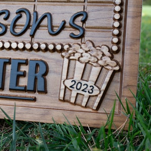 Pu&ograve; includere: Un cartello in legno con il testo "Wilson's Theater" e un secchiello di popcorn intagliato con l'anno "2023" sopra.