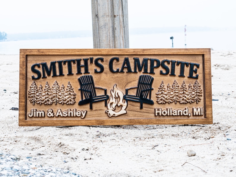 Puede incluir: Un letrero de madera con el texto "Smith's Campsite" en letras negras. El letrero tambi&eacute;n presenta dos sillas Adirondack negras, una fogata y una hilera de pinos a cada lado. El texto "Jim & Ashley" aparece en el lado izquierdo del letrero, y "Holland, MI" aparece en el lado derecho.