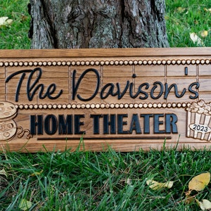 Pu&ograve; includere: Un cartello in legno con il testo "The Davison's Home Theater" in lettere nere. Il cartello &egrave; decorato con una bobina di pellicola e un secchio di popcorn con l'anno 2023 sopra.