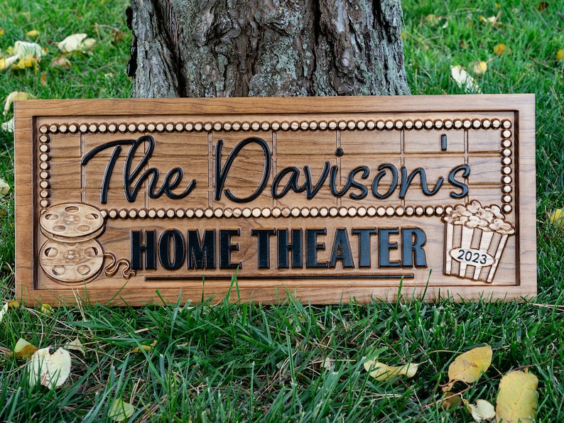 Pu&ograve; includere: Un cartello in legno con il testo "The Davison's Home Theater" in lettere nere. Il cartello &egrave; decorato con una bobina di pellicola e un secchio di popcorn con l'anno 2023 sopra.