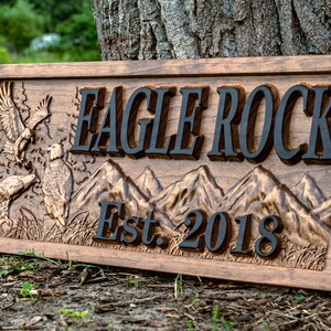 May include: A wooden sign with the words "Eagle Rock Est. 2018" carved into it. The sign features a carved image of mountains and eagles.