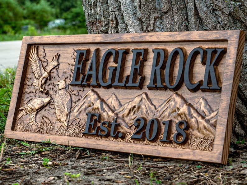 May include: A wooden sign with the words "Eagle Rock Est. 2018" carved into it. The sign features a carved image of mountains and eagles.