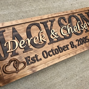 May include: A wooden sign with the names "Jackson" and "Chelsey" in a dark brown font, with "Derek & Chelsey" in a lighter font. The sign also includes "Est. October 8, 2005" and two carved hearts.