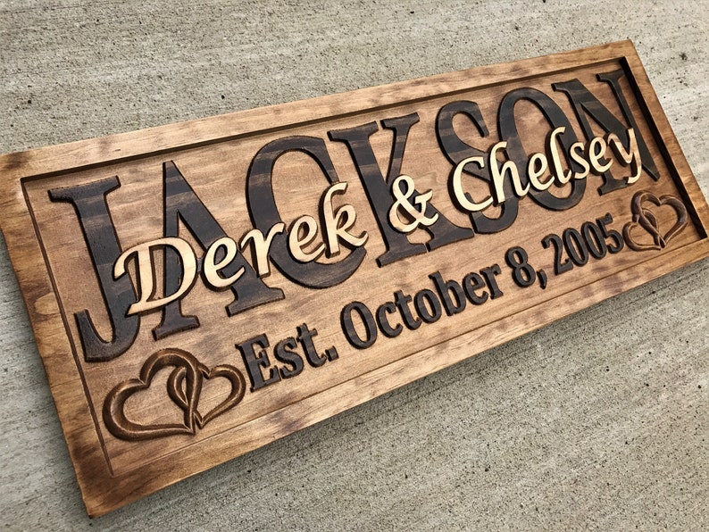 May include: A wooden sign with the names "Jackson" and "Chelsey" in a dark brown font, with "Derek & Chelsey" in a lighter font. The sign also includes "Est. October 8, 2005" and two carved hearts.
