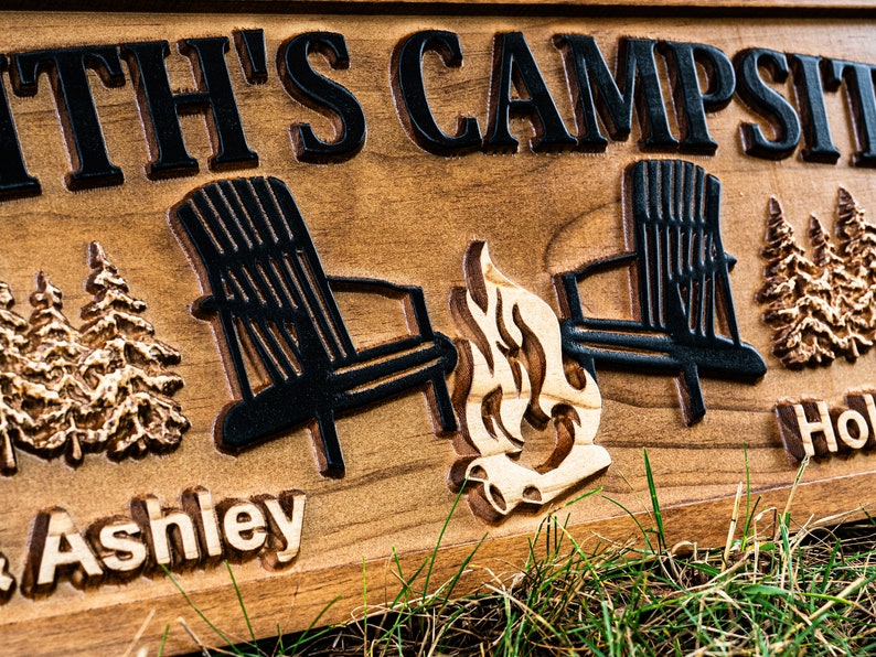 Puede incluir: Un letrero de madera con el texto "Smith's Campsite" y los nombres "Ashley" y "Hol", con una imagen tallada de dos sillas Adirondack, una fogata y pinos.