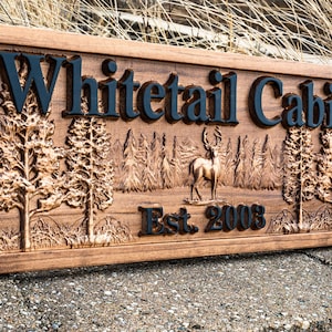 Peut inclure: Une pancarte en bois avec le texte "Whitetail Cabin Est. 2003" grav&eacute; dessus. La pancarte pr&eacute;sente une image grav&eacute;e d'un cerf dans un d&eacute;cor de for&ecirc;t.