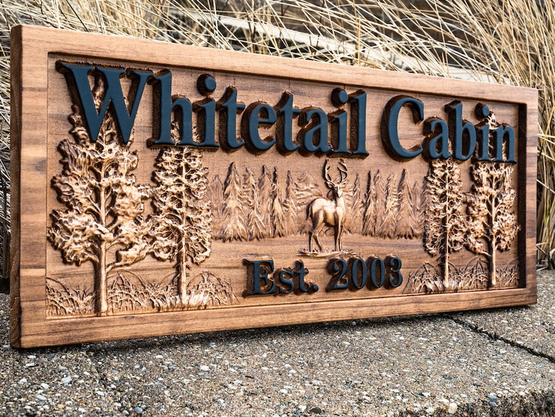 Peut inclure: Une pancarte en bois avec le texte "Whitetail Cabin Est. 2003" grav&eacute; dessus. La pancarte pr&eacute;sente une image grav&eacute;e d'un cerf dans un d&eacute;cor de for&ecirc;t.