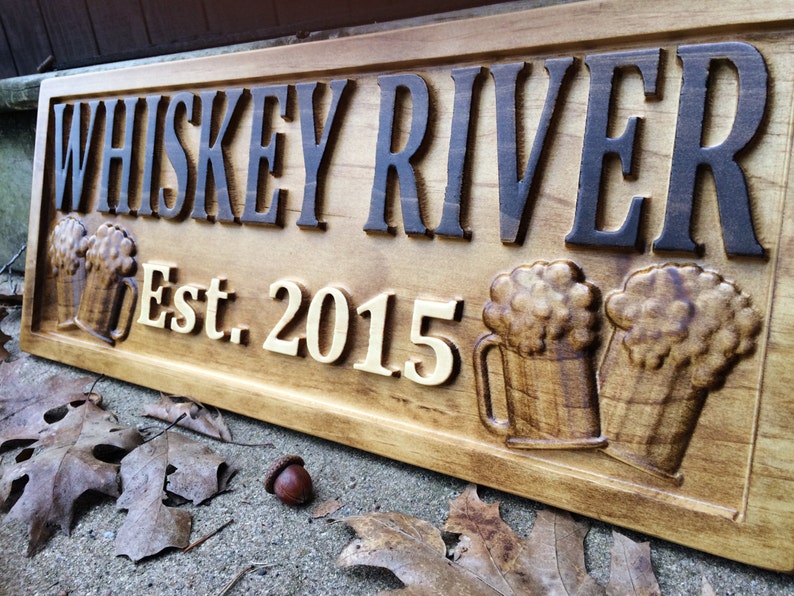 May include: Wooden sign with the text "Whiskey River Est. 2015" in brown and gold lettering. The sign features two carved wooden beer mugs with foam on top.