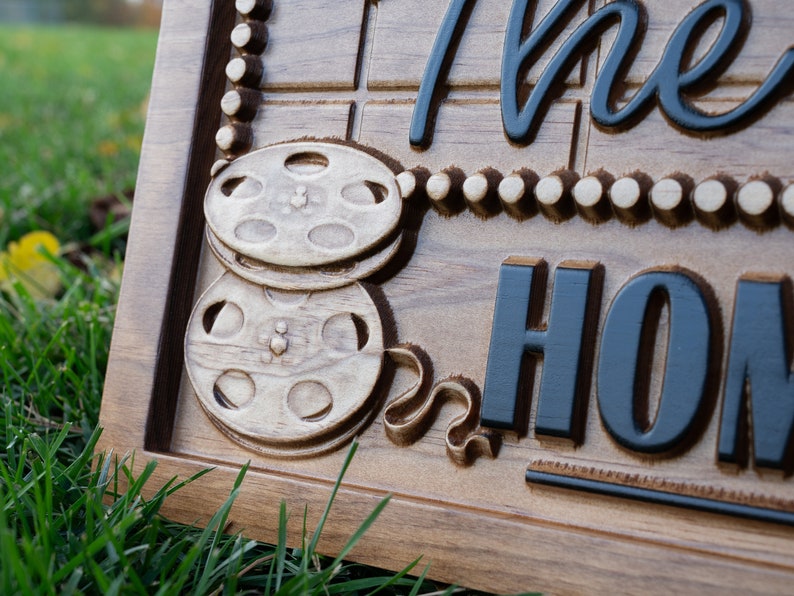 Pu&ograve; includere: Un cartello in legno con le parole "The Home" in lettere nere. Il cartello presenta una bobina di pellicola con un nastro nero e una fila di perline bianche.