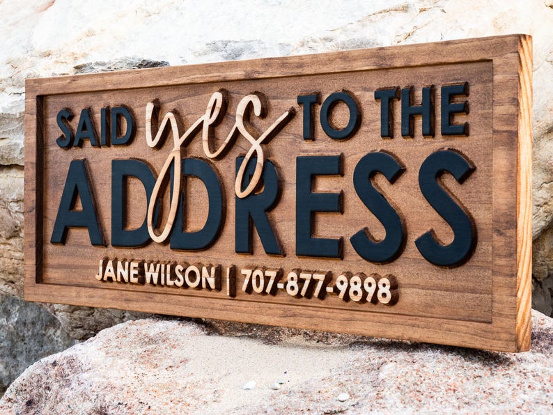 May include: A rectangular wooden sign with the words "SAID yes TO THE ADDRESS" in a layered design. The word "yes" is in a cursive font. The name "JANE WILSON" and a phone number are at the bottom.