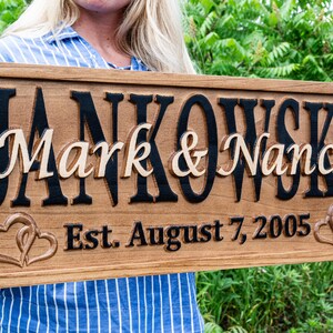 May include: Wooden sign with the names "Mark & Nancy" in a light colour, set against a dark background. The sign also includes the date "Est. August 7, 2005" and two intertwined hearts.