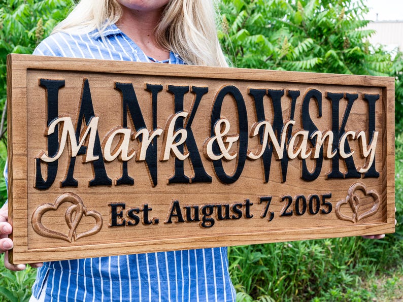 May include: Wooden sign with the names "Mark & Nancy" in a light colour, set against a dark background. The sign also includes the date "Est. August 7, 2005" and two intertwined hearts.