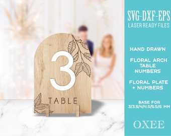 May include: A laser-cut wooden table number with a floral arch design. The number 3 is displayed in white on a brown wooden background. The text "TABLE" is below the number. The image includes the text "SVG-DXF-EPS LASER READY FILES", "HAND DRAWN", "FLORAL ARCH TABLE NUMBERS", "FLORAL PLATE + NUMBERS", "BASE FOR 3/3.5/4/4.5/5.5/6 MM", and "OXEE".