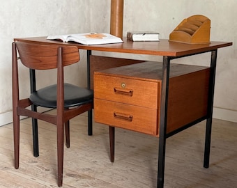Op de afbeelding: Een houten bureau met een zwart metalen frame en twee lades, vergezeld door een bijpassende houten stoel met een zwarte zitting. Op het bureaublad liggen een open boek, een gesloten boek, een hoge bruine vaas en een houten brievenhouder.