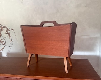 Op de afbeelding: Een houten tijdschriftenrek met een warme bruine nerf, een rechthoekig lichaam met een centrale uitgesneden handgreep aan de bovenkant en vier schuine, taps toelopende poten. De bovenkant is open voor het opbergen van tijdschriften.