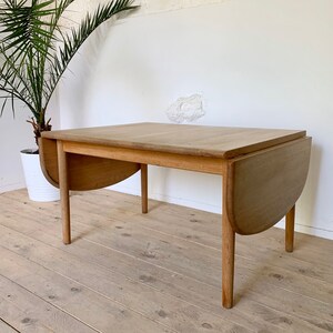 Puede incluir: Una mesa ovalada de madera clara con hojas abatibles y acabado natural. La mesa tiene cuatro patas y est&aacute; hecha de madera maciza. La mesa est&aacute; en una habitaci&oacute;n con suelo de madera clara.