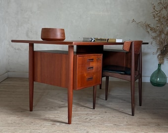 Op de afbeelding: Een houten bureau met een rechthoekig blad, vier taps toelopende poten en een zijkast met drie gestapelde lades. Een bijpassende houten stoel met een zwarte zitting staat eronder. Op het bureau staan een houten kom en enkele boeken.