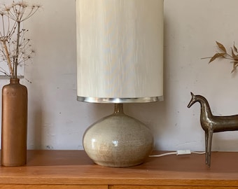 Op de afbeelding: Een keramische tafellamp met een crèmekleurige lampenkap en een zilveren rand. De lamp heeft een afgeronde voet en staat op een houten oppervlak. Een bronzen beeld van een hert en een bruine fles met gedroogde bloemen zijn ook aanwezig.