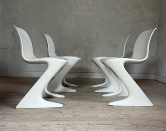 Op de afbeelding: Een set van vier witte cantilever stoelen, elk met een doorlopende, enkelvoudige gegoten vorm met een gebogen rugleuning en een Z-vormige basis die uitloopt in een platte, stabiele voet, opgesteld in twee paren.