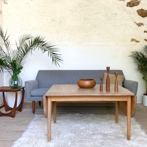 Puede incluir: Un sof&aacute; gris claro con una mesa de centro de madera delante. La mesa de centro tiene un cuenco de madera y tres candelabros de madera en la parte superior. El sof&aacute; est&aacute; en una habitaci&oacute;n con paredes blancas y una alfombra en el suelo.