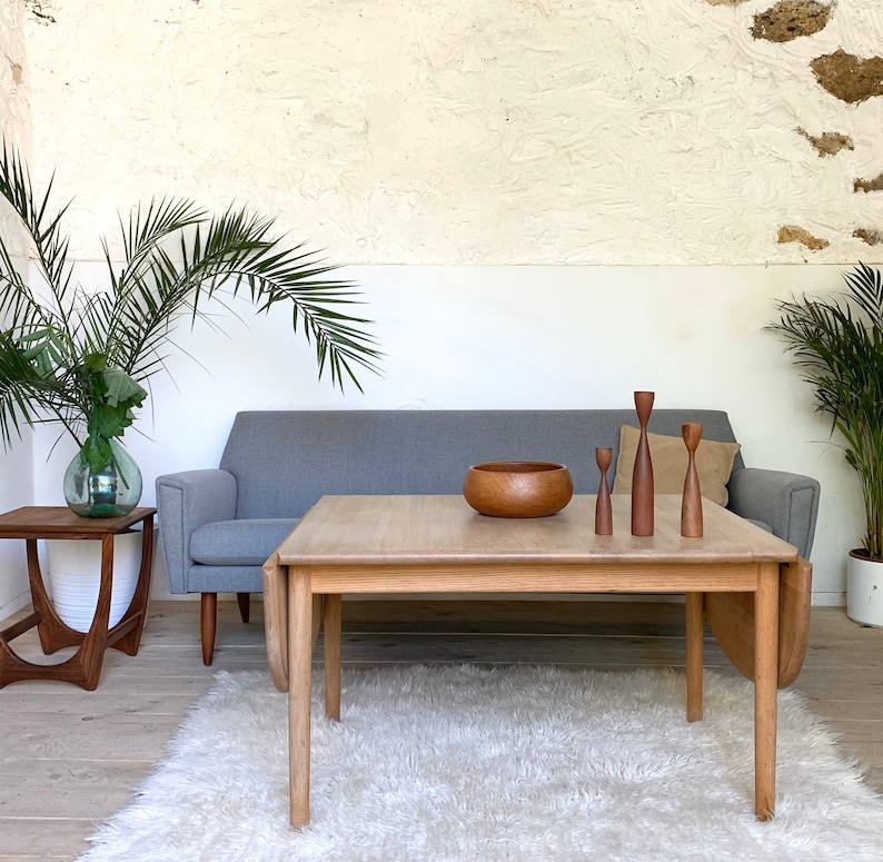 Puede incluir: Un sof&aacute; gris claro con una mesa de centro de madera delante. La mesa de centro tiene un cuenco de madera y tres candelabros de madera en la parte superior. El sof&aacute; est&aacute; en una habitaci&oacute;n con paredes blancas y una alfombra en el suelo.