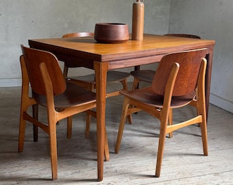 Op de afbeelding: Een rechthoekige houten eettafel met vier bijpassende stoelen. De tafel heeft een glad, middelbruin houten oppervlak met een zichtbare nerf. Elke stoel heeft een gebogen houten rugleuning en vier taps toelopende poten. Een houten kom en vaas.