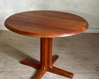 Op de afbeelding: Een ronde houten tafel met een warme bruine afwerking, voorzien van een glad cirkelvormig blad en een centrale kolomvoet die uitloopt in vier stevige, gebogen poten. De houtnerf is zichtbaar over het gehele oppervlak en toont een natuurlijke textuur.