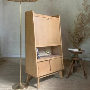 Peut inclure: Un meuble en bois clair avec une porte rabattable et deux portes inférieures. Une figurine d'oiseau en laiton est posée sur une étagère à l'intérieur. Un lampadaire avec un abat-jour tissé et un petit tabouret en bois sont également dans la pièce.