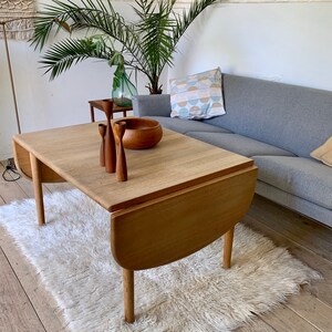 Puede incluir: Una mesa de comedor extensible de madera clara con un borde redondeado y una alfombra blanca y esponjosa debajo. La mesa tiene un acabado de madera natural y est&aacute; puesta con tres candelabros de madera y un cuenco de madera.