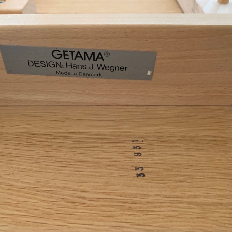 Puede incluir: Un primer plano de un caj&oacute;n de madera con una etiqueta que dice "GETAMA&reg; DESIGN: Hans J. Wegner Hecho en Dinamarca". El caj&oacute;n tambi&eacute;n tiene un n&uacute;mero "53 931" estampado.