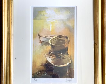 Op de afbeelding: Een ingelijste kunstprint met drie kleine boten op het water, in een goudkleurige lijst. De boten zijn bruin en wit, tegen een zachte gele en beige achtergrond. De print is genummerd 2/250 en gesigneerd.