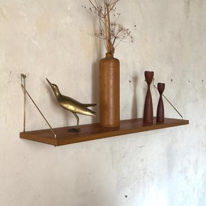May include: A wooden shelf with brass supports displays a brass bird sculpture, a brown ceramic bottle with dried flowers, and two wooden candle holders. The shelf is mounted on a textured, off-white wall, creating a vintage aesthetic.