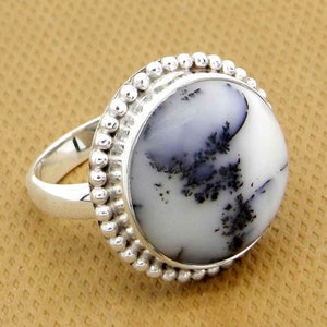 May include: A silver ring featuring a large, round, white gemstone with black dendritic inclusions. The gemstone is surrounded by a beaded silver border. The ring has a simple, polished silver band.