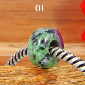 Könnte beinhalten: Eine grüne und violette Edelsteinperle mit schwarzen Flecken auf einer silbernen Kette. Die Perle ist rund mit einem Loch in der Mitte. Die Perle liegt auf einer Holzoberfläche. 01