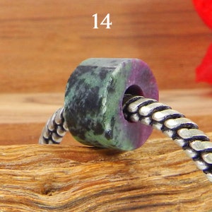 May include: A cylindrical gemstone bead with a hole, showing green, black, and purple colours. The bead is strung on a silver-coloured chain. The number "14" is visible in the upper left corner.