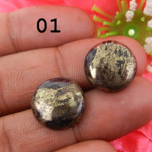 Puede incluir: Dos cabujones redondos de pirita negra y dorada. Los cabujones tienen una superficie lisa y pulida y un patrón natural irregular de inclusiones de pirita dorada.