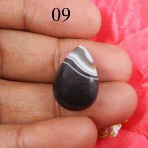 May include: Black and white agate cabochon in a teardrop shape. The stone has a layered pattern of white and black bands.