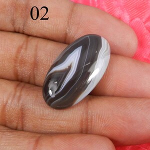 May include: A black and white banded agate cabochon with a smooth, polished surface. The cabochon is oval shaped and has a natural, organic pattern.