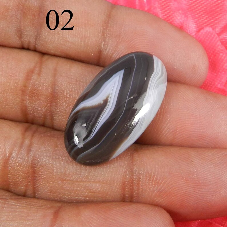 May include: A black and white banded agate cabochon with a smooth, polished surface. The cabochon is oval shaped and has a natural, organic pattern.