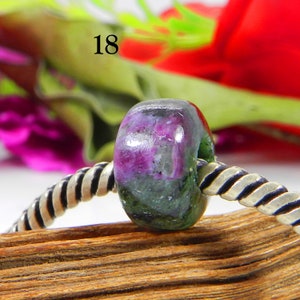 Könnte beinhalten: Eine grüne und rosafarbene Edelsteinperle mit einem silbernen und schwarzen geflochtenen Armband. Die Perle hat einen Durchmesser von etwa 1 cm. Die Perle ist ein natürlicher Edelstein und hat ein einzigartiges Muster aus Grün und Rosa.