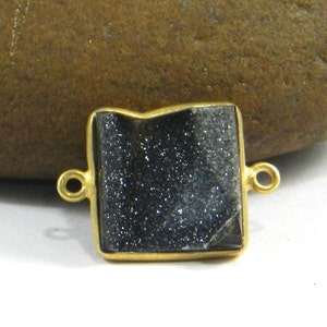 Pode incluir: Conector folheado a ouro com uma pedra preciosa preta quadrada. A pedra preciosa tem uma textura brilhante e cintilante.
