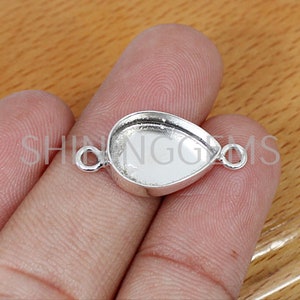 May include: A silver-colored teardrop-shaped jewelry finding with a bezel setting and two small loops. The finding is designed to hold a gemstone or other decorative element. The text "SHINGGEMS" is visible.