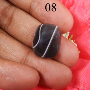 May include: A black and white agate cabochon with a curved shape. The stone has a dark gray base with white stripes.